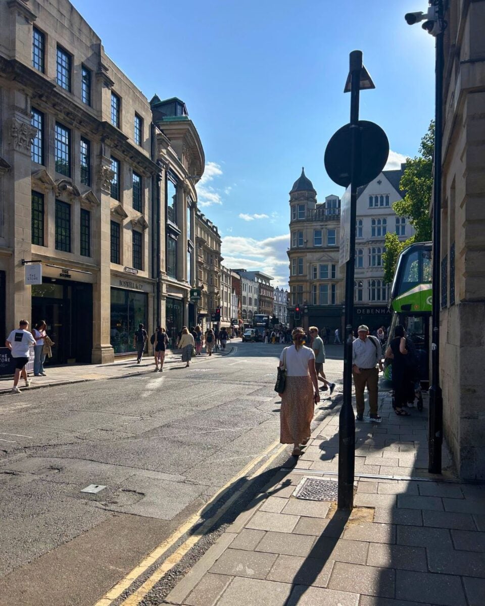 Sunny city street scene with pedestrians walking along sidewalks and historic buildings lining the road under a clear blue sky.