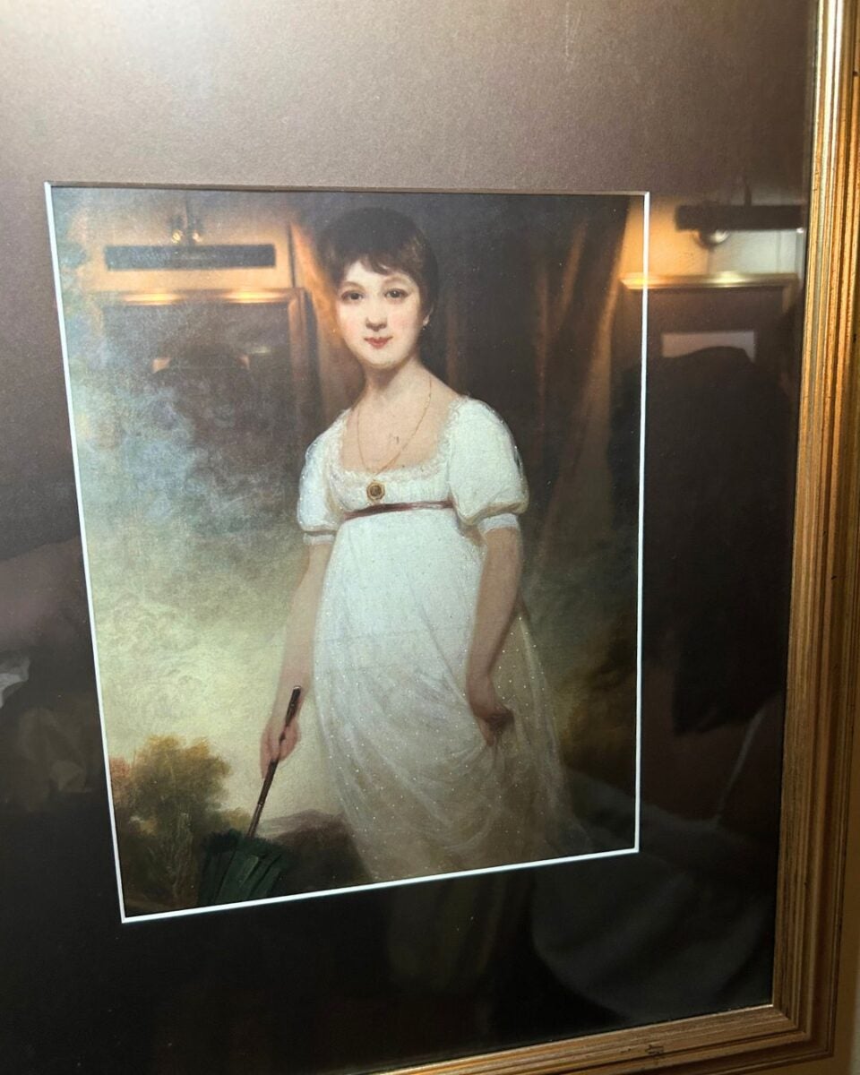 Portrait of a young woman in a white empire-waist dress holding a fan, set against a softly lit background with warm tones.