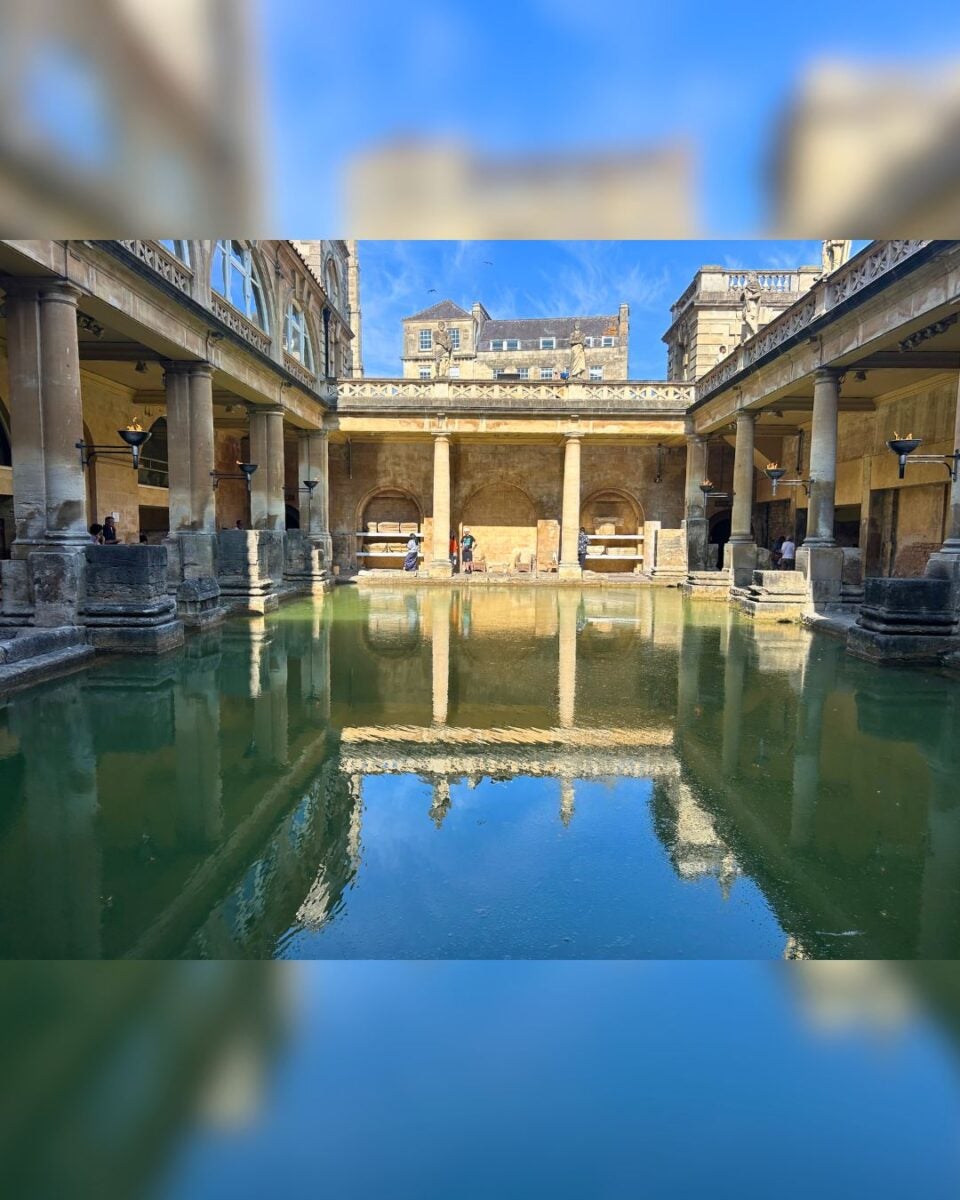Historic Roman-style bathhouse with stone columns surrounding a reflective pool under a clear blue sky.