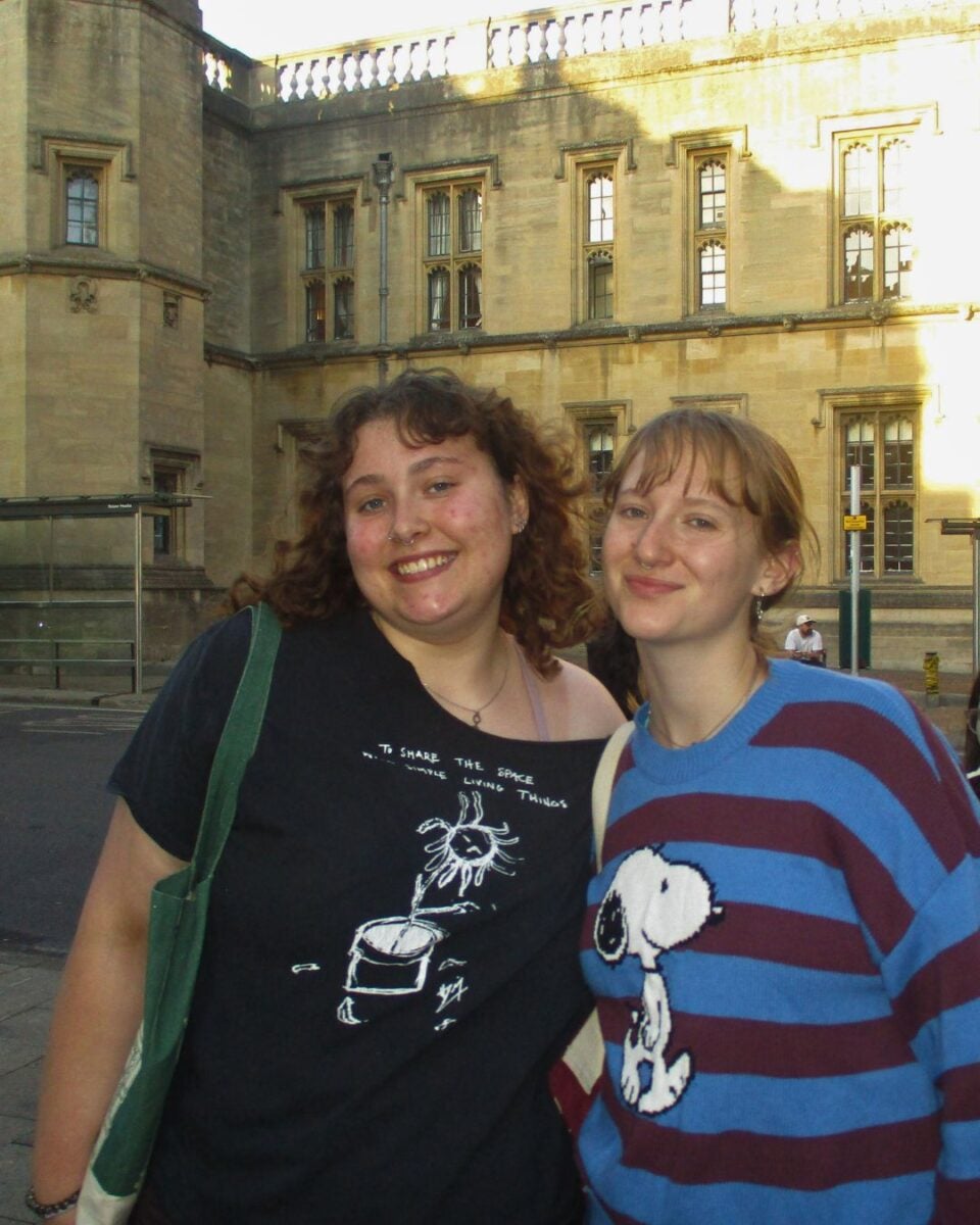 Two young women smiling outdoors in front of a historic stone building with large windows on a sunny day.