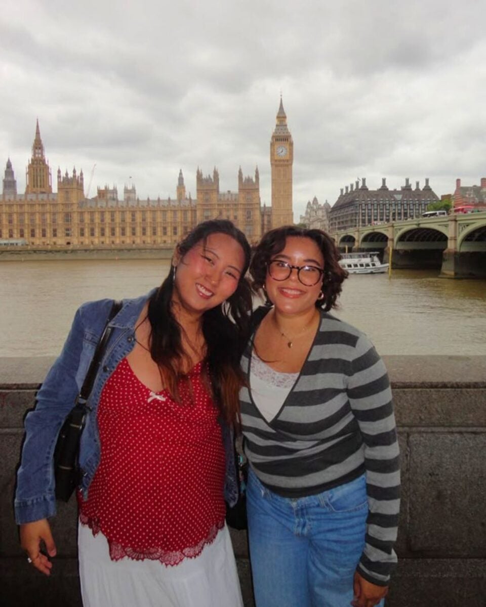 Two women smiling by the River Thames with the Houses of Parliament and Big Ben in the background on a cloudy day.