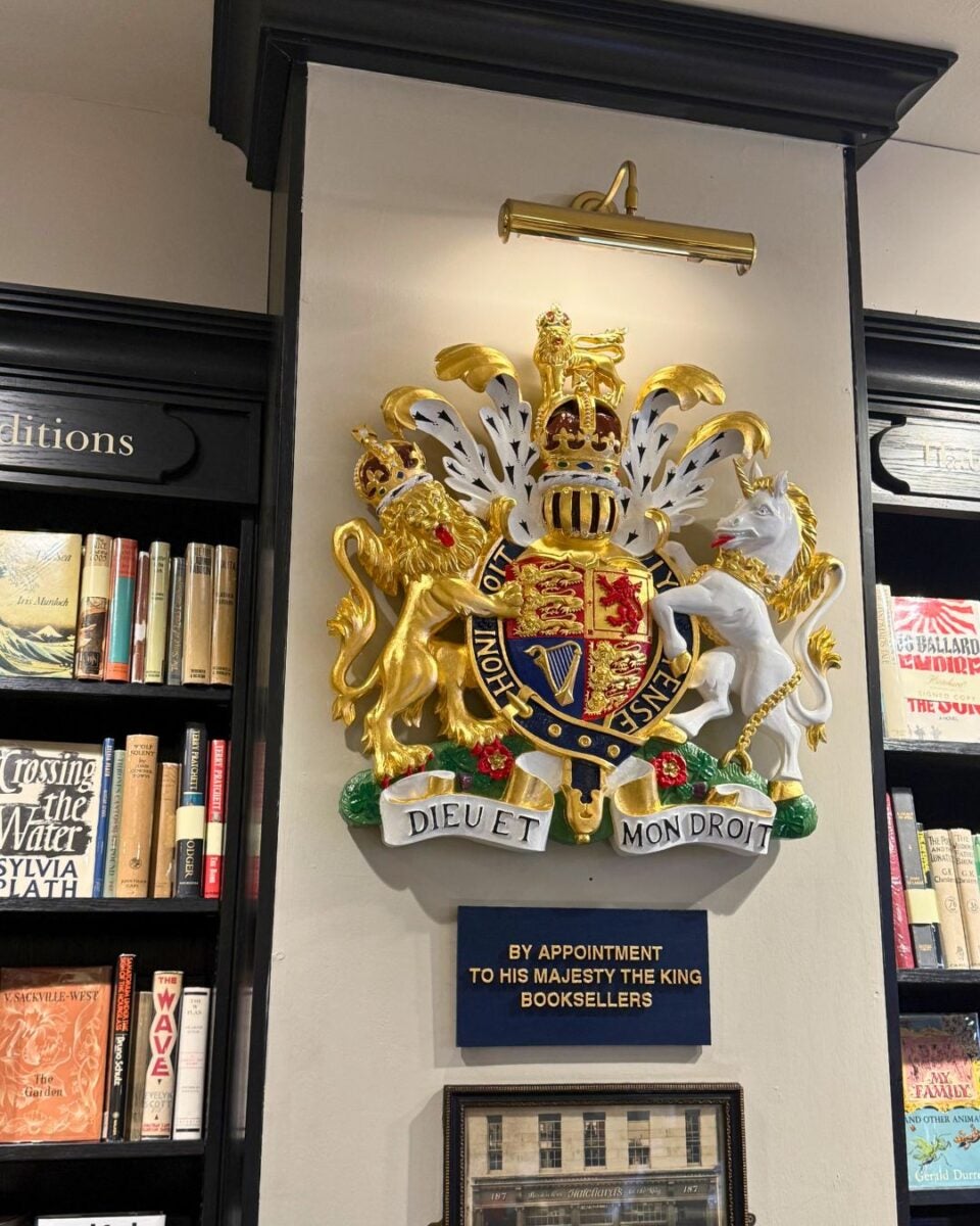 Royal coat of arms featuring a lion and unicorn above a plaque reading 'By Appointment to His Majesty The King Booksellers' in a bookstore.