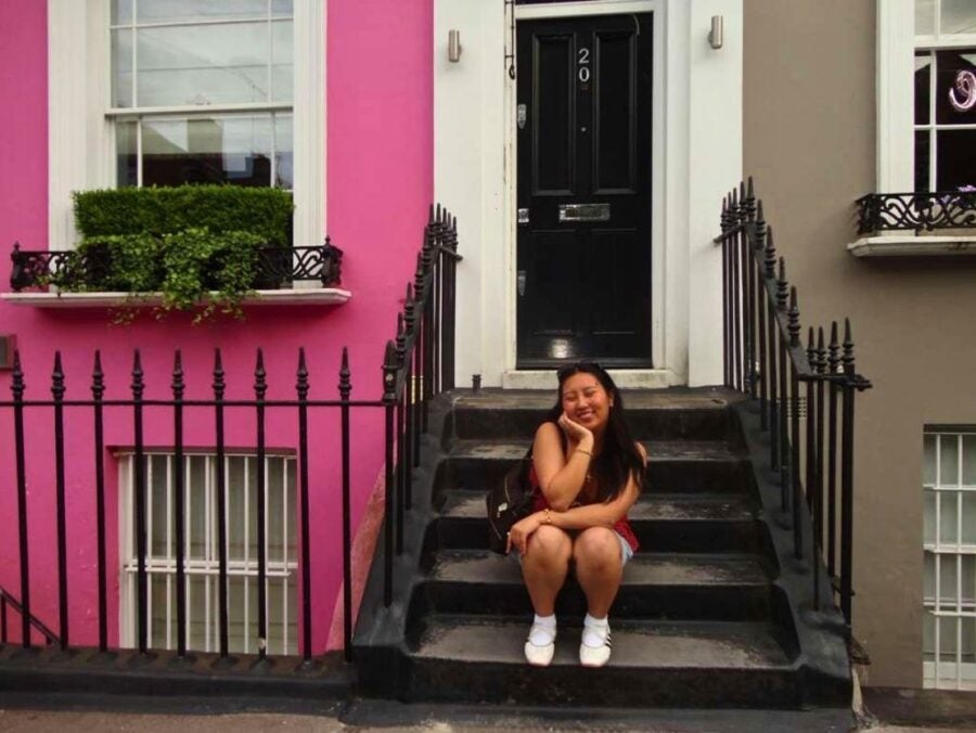 Young woman sitting on black stone steps outside a black door numbered 20, smiling with eyes closed and resting her head on her hand.