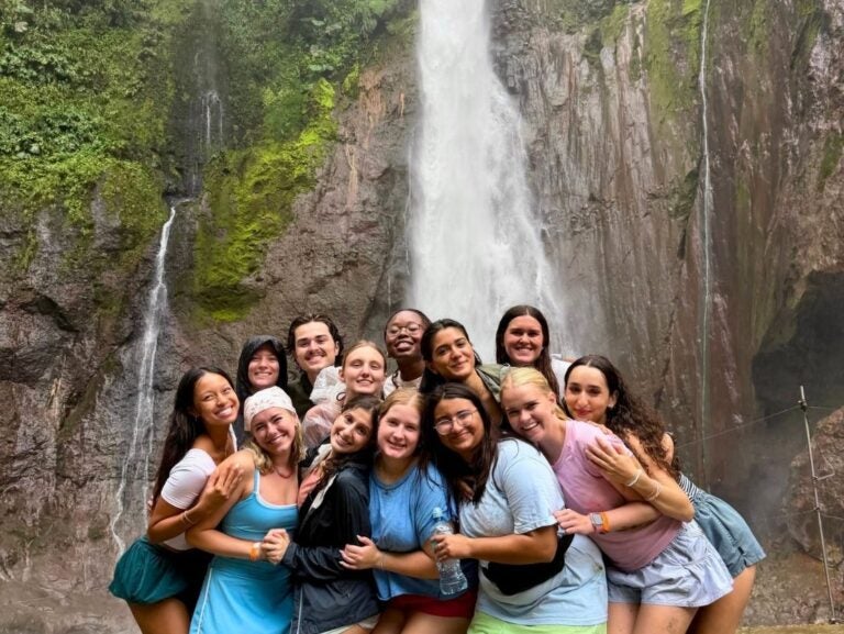 Group of thirteen friends smiling and hugging in front of a tall waterfall surrounded by lush greenery and rocky cliffs.