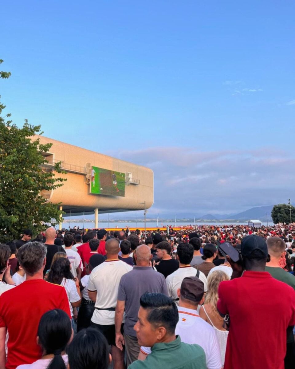Large crowd gathered outdoors watching a soccer match on a big screen mounted on a modern building at dusk.