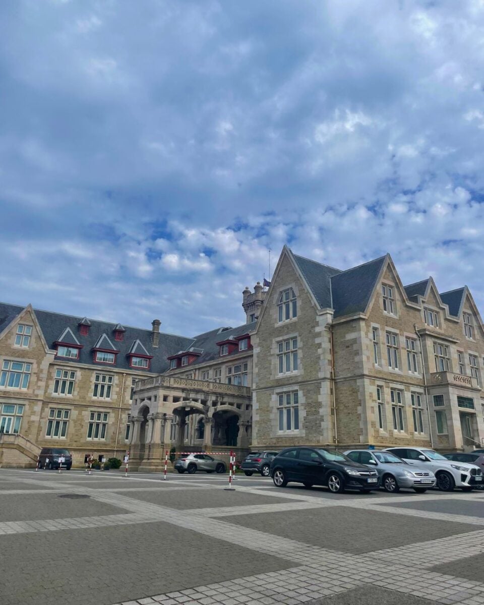 Large historic stone building with steep gabled roofs under a cloudy sky, surrounded by parked cars in a spacious courtyard.