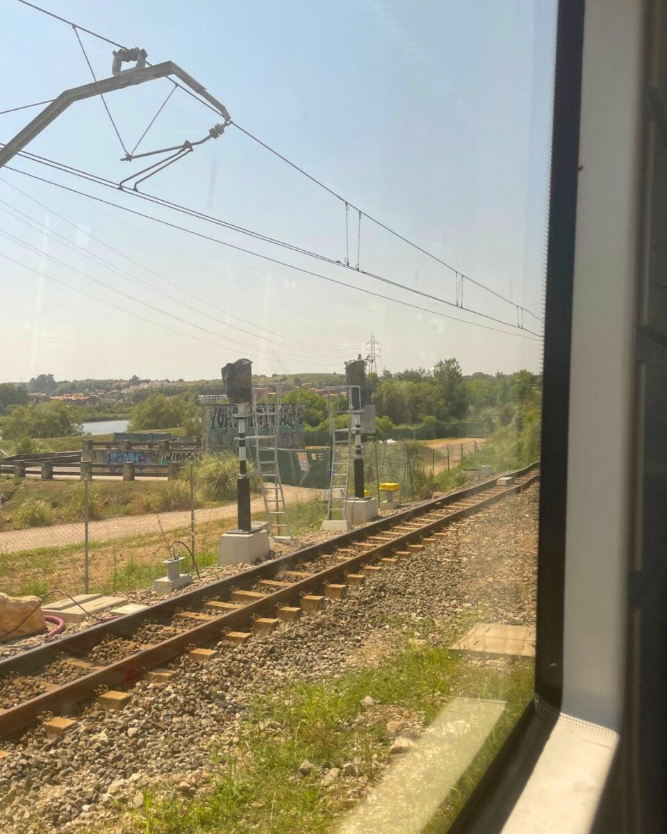 View of railway tracks and overhead electric lines seen through a train window on a clear day.