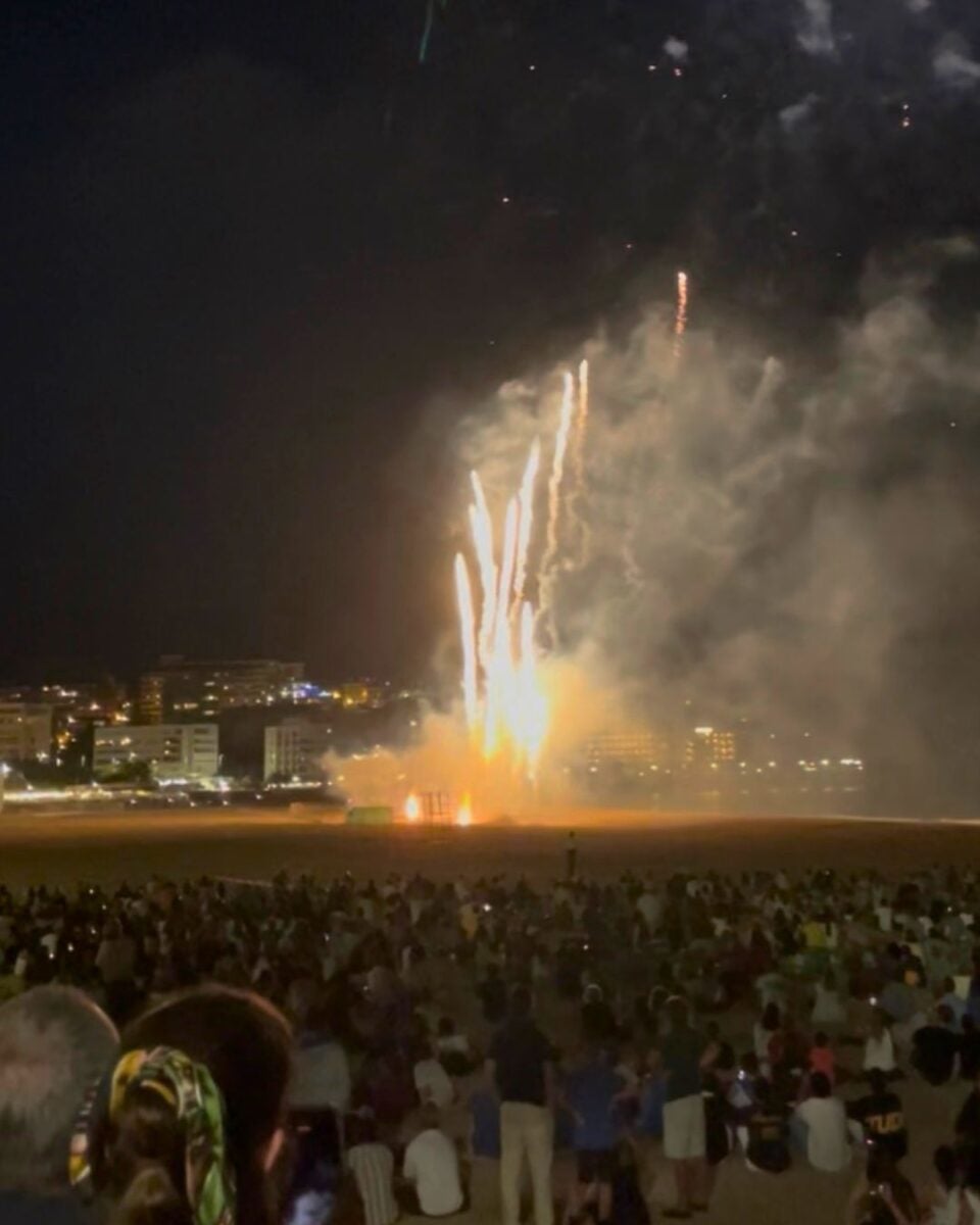 Crowd gathered on a beach at night watching bright fireworks shooting into the sky with city lights in the background.