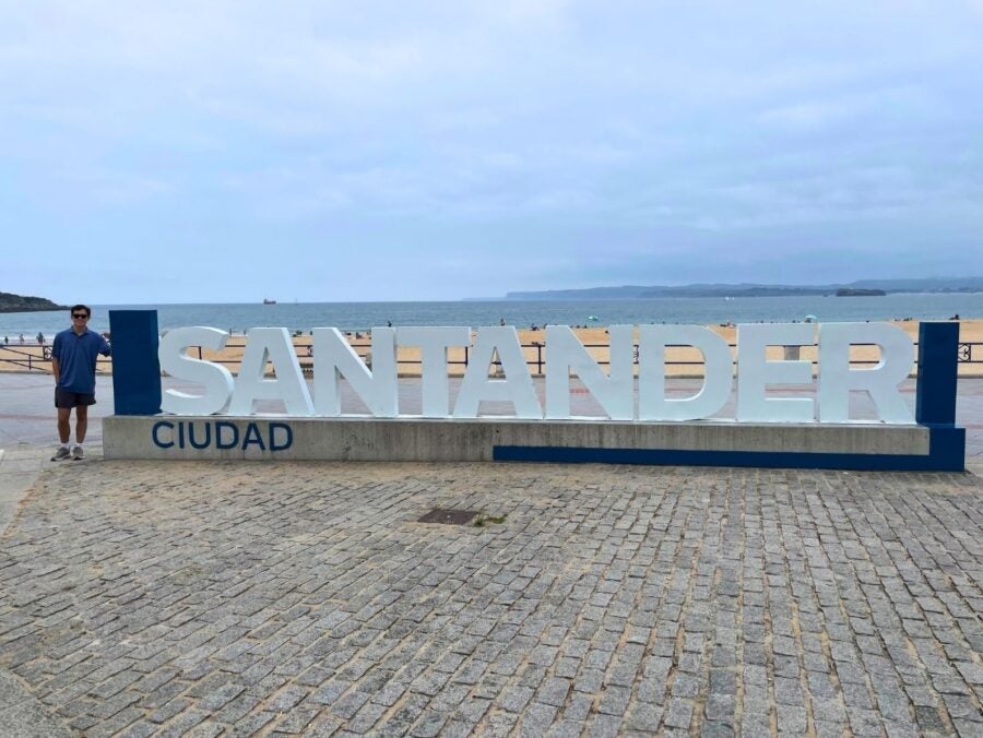 Large white letters spelling 'SANTANDER' with 'CIUDAD' below, set on a stone plaza by the waterfront under a cloudy sky.