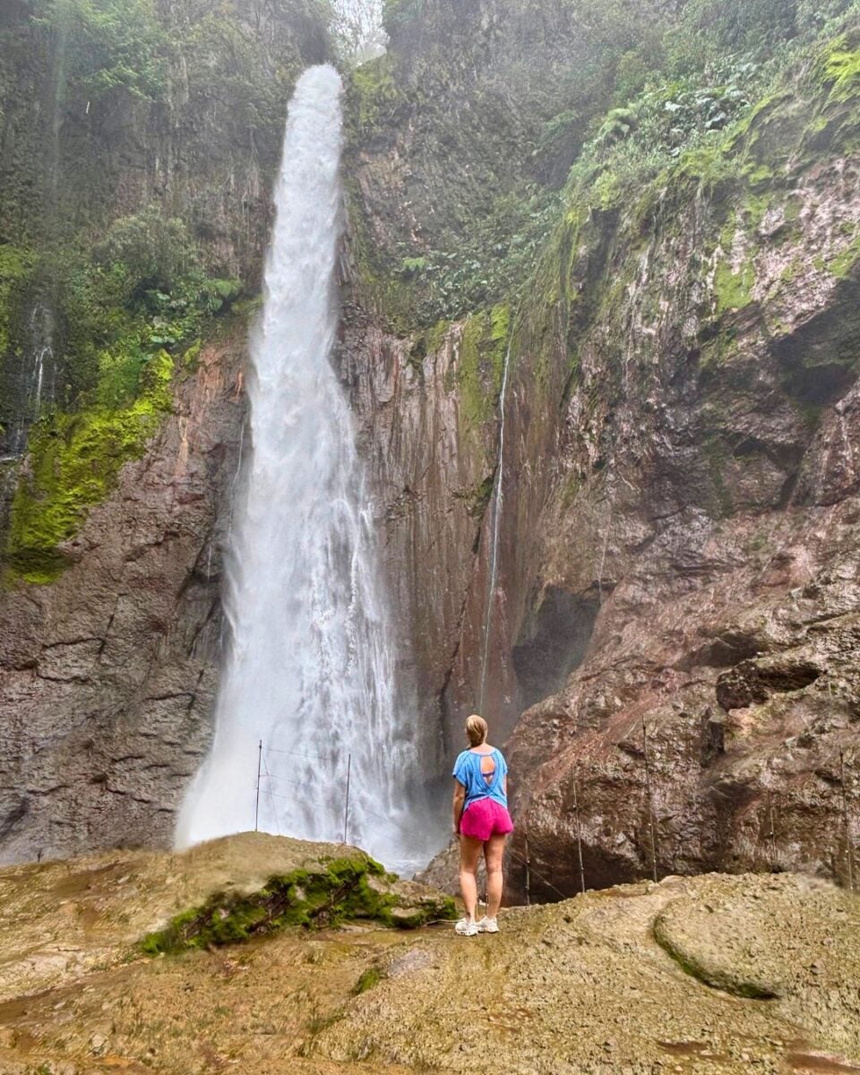 A woman in pink shorts and a blue shirt stands on rocky ground, gazing up at a tall, powerful waterfall surrounded by lush greenery and steep cliffs.