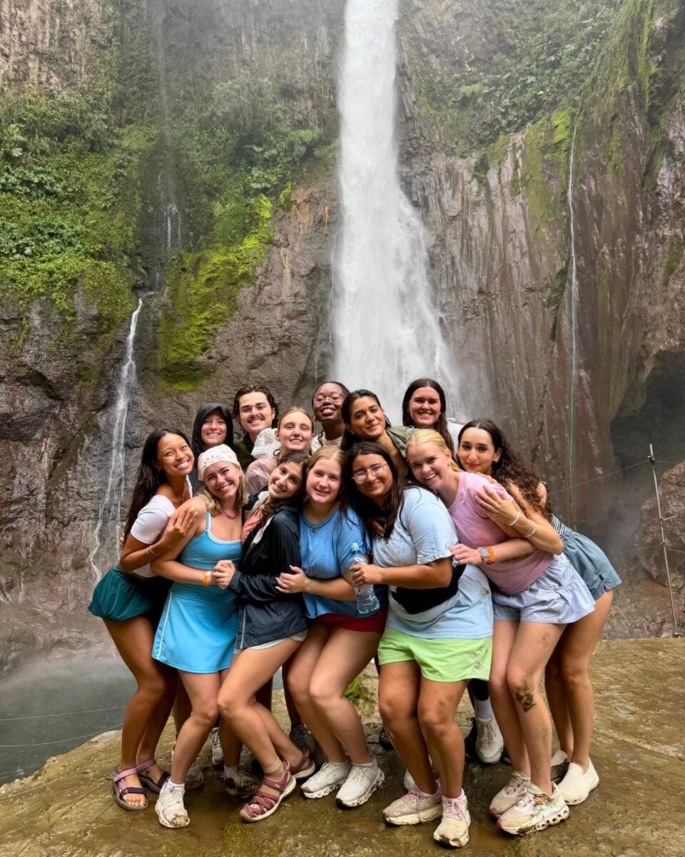 Group of thirteen friends smiling and hugging in front of a tall waterfall surrounded by lush greenery and rocky cliffs.