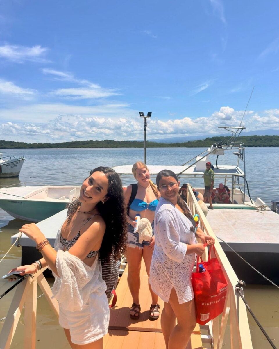 Three women in summer attire smiling on a wooden dock with boats and calm water under a blue sky.