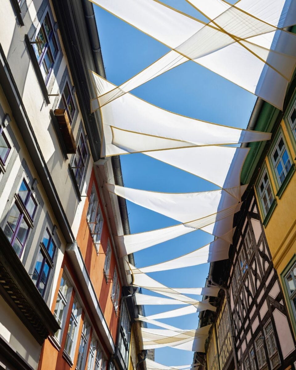 White triangular fabric shades stretch across a narrow street between colorful historic buildings under a clear blue sky.