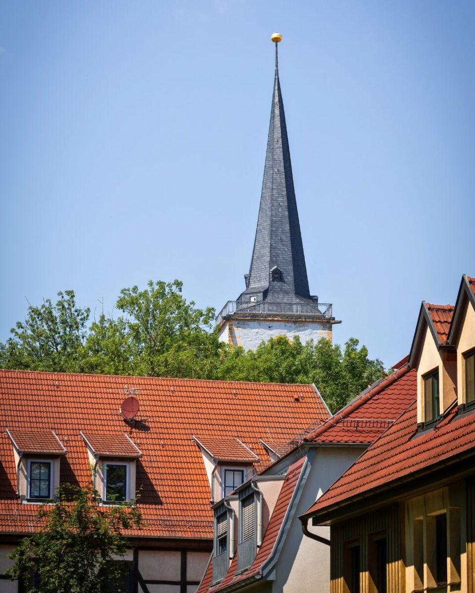 Tall church steeple with a golden sphere atop rising above green trees and red-tiled rooftops under a clear blue sky.