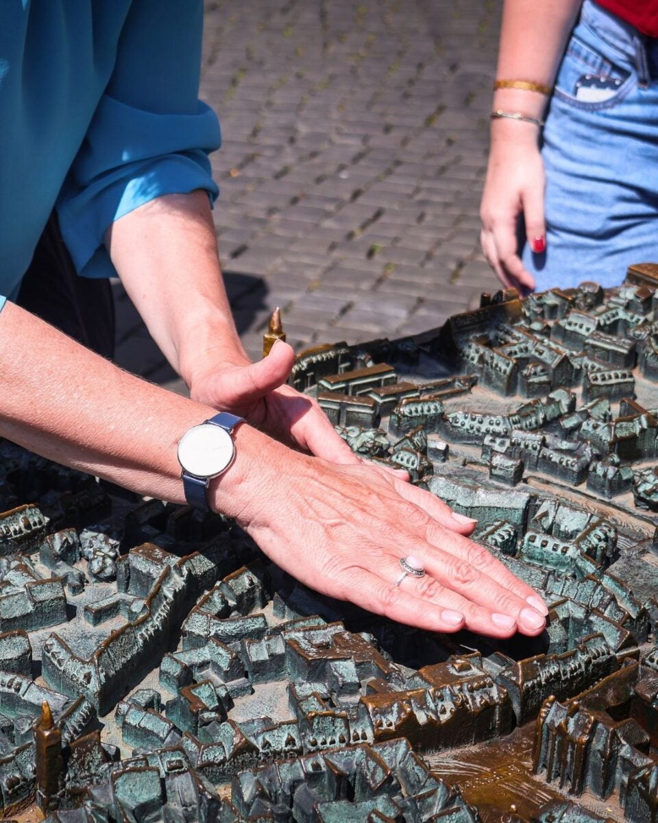 Person's hands exploring a detailed bronze tactile model of a historic city layout on a cobblestone street.
