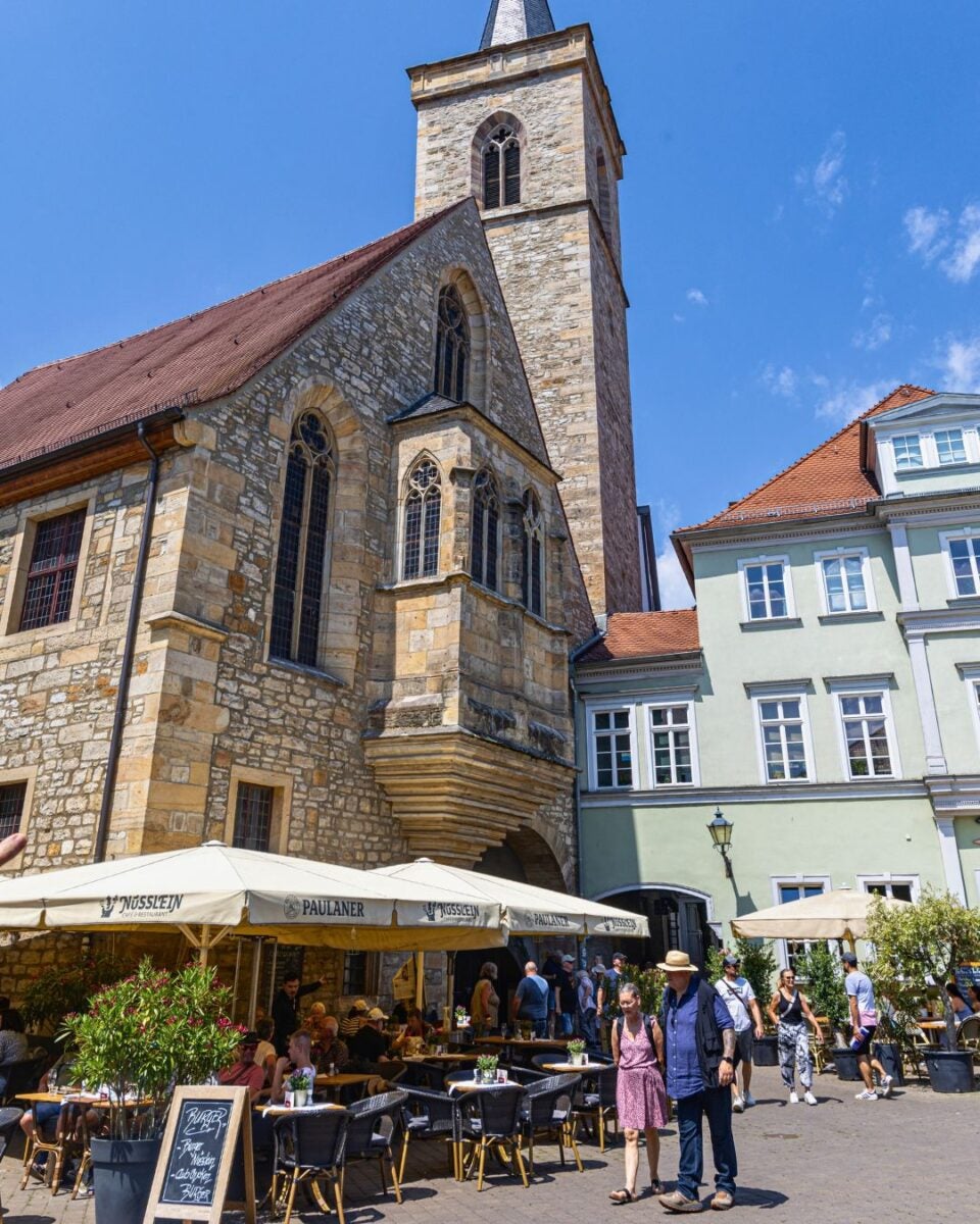 Historic stone church with tall tower overlooking a bustling outdoor café filled with people on a sunny day.