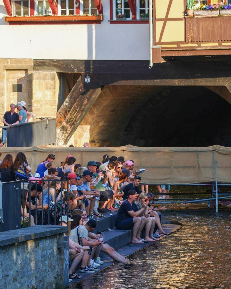 People sitting on stone steps by a river under a historic arched bridge, enjoying the sunny day and water views.