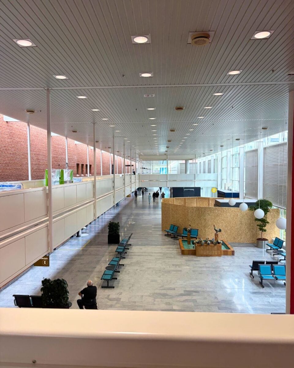 Spacious, modern airport terminal with rows of blue seating, high ceiling lights, and a few travelers walking in the distance.
