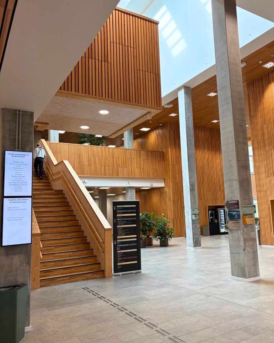 Modern interior of a building featuring wooden panel walls, a wide staircase, and large concrete pillars under a skylight ceiling.