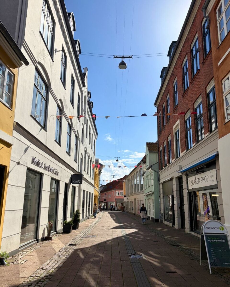 Sunny narrow street lined with colorful historic buildings and small shops under a clear blue sky with hanging streetlights and festive bunting.