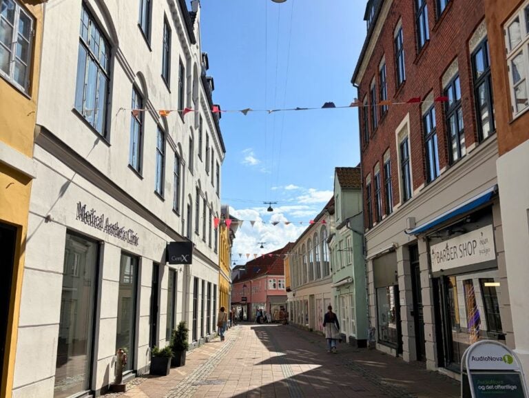 Sunny narrow street lined with colorful historic buildings and small shops under a clear blue sky with hanging streetlights and festive bunting.