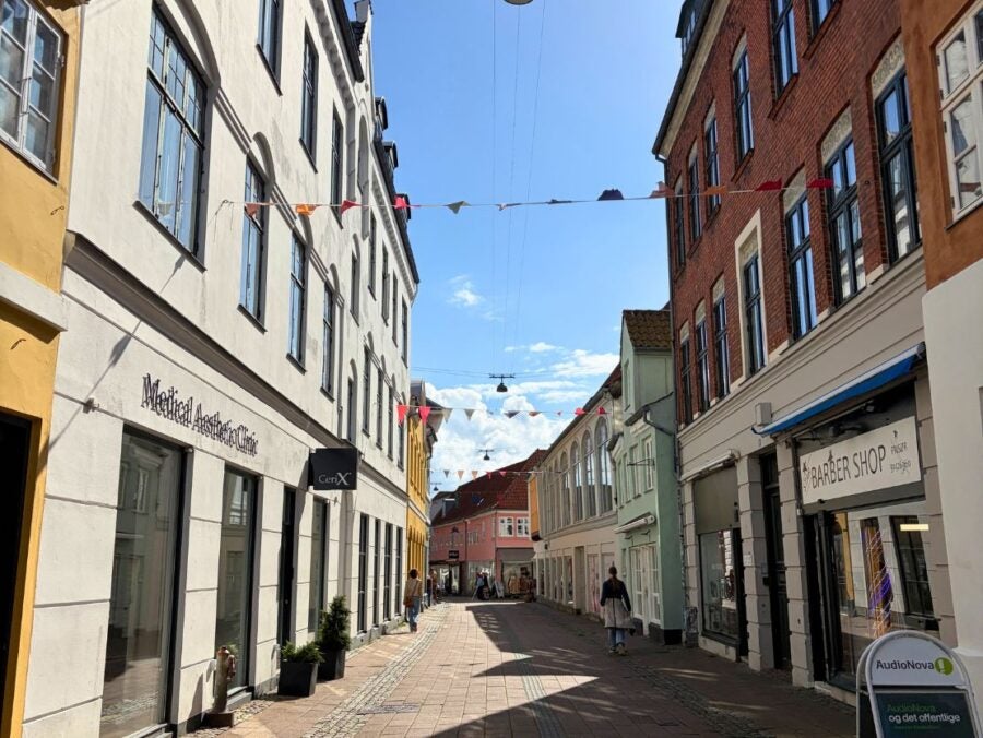 Sunny narrow street lined with colorful historic buildings and small shops under a clear blue sky with hanging streetlights and festive bunting.