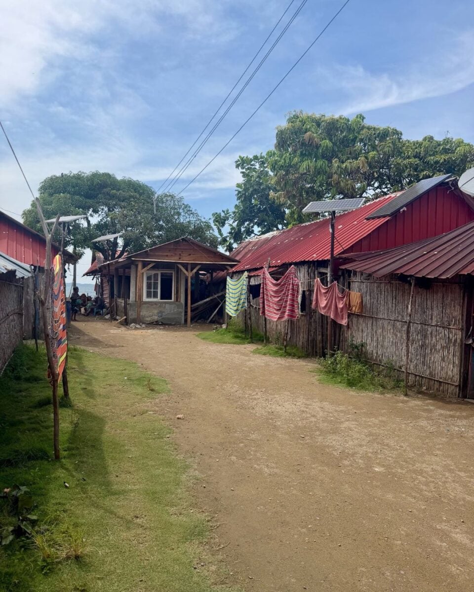 Dirt road flanked by rustic houses with red metal roofs and clothes hanging on bamboo fences under a partly cloudy sky.