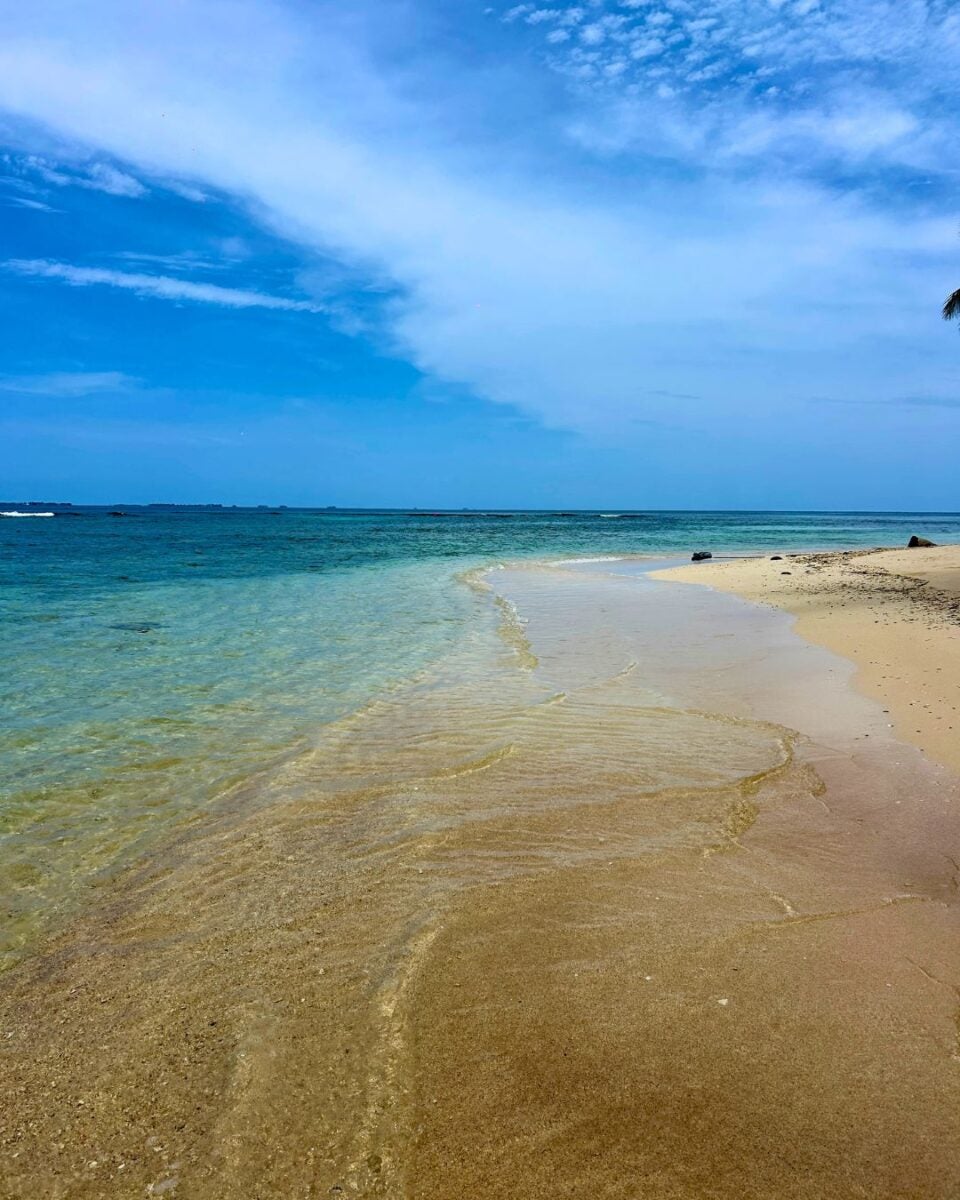 Clear turquoise water gently laps onto a sandy beach under a partly cloudy blue sky.