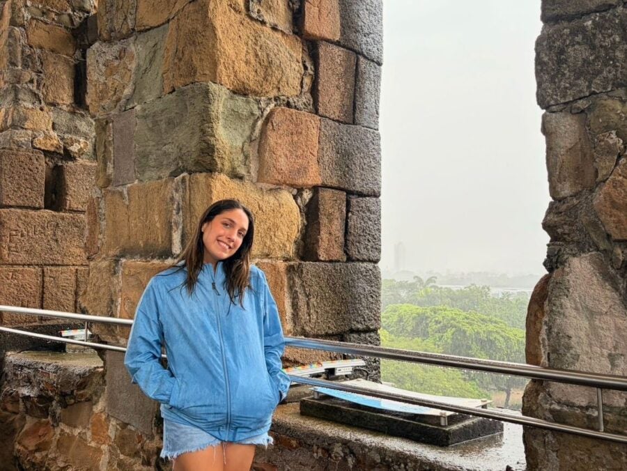 Young woman in a blue jacket and denim shorts leaning against a stone wall with a scenic green landscape in the background.