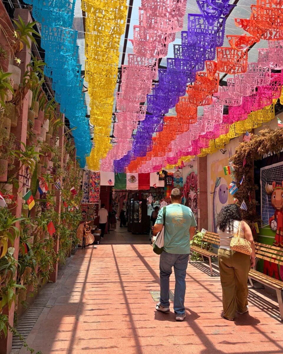 Colorful papel picado banners hang overhead in a vibrant outdoor corridor with people walking beneath them.