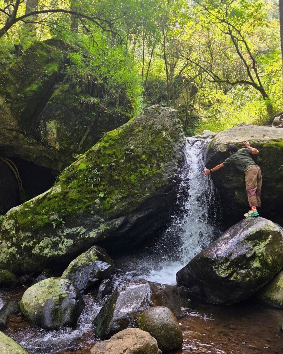 A young man in outdoor gear reaching toward a small waterfall surrounded by moss-covered rocks and lush green forest.