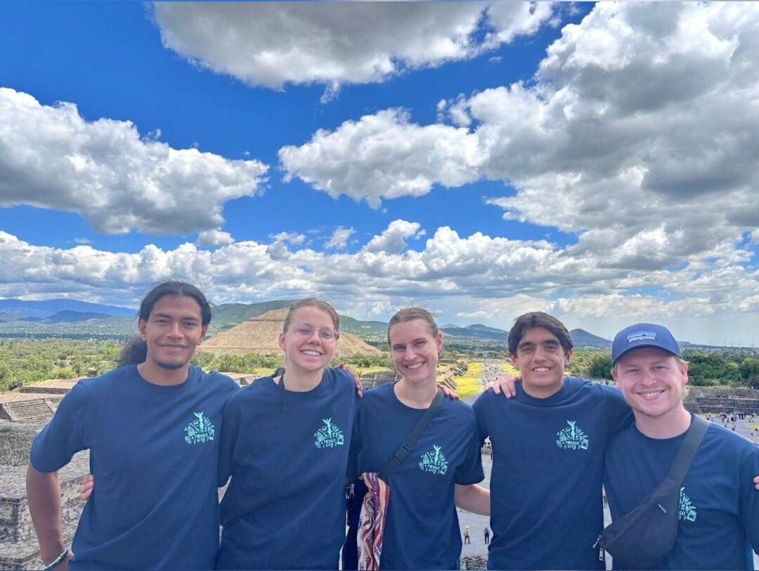 Group of five young adults smiling and posing together outdoors under a bright blue sky with scattered clouds and distant hills.