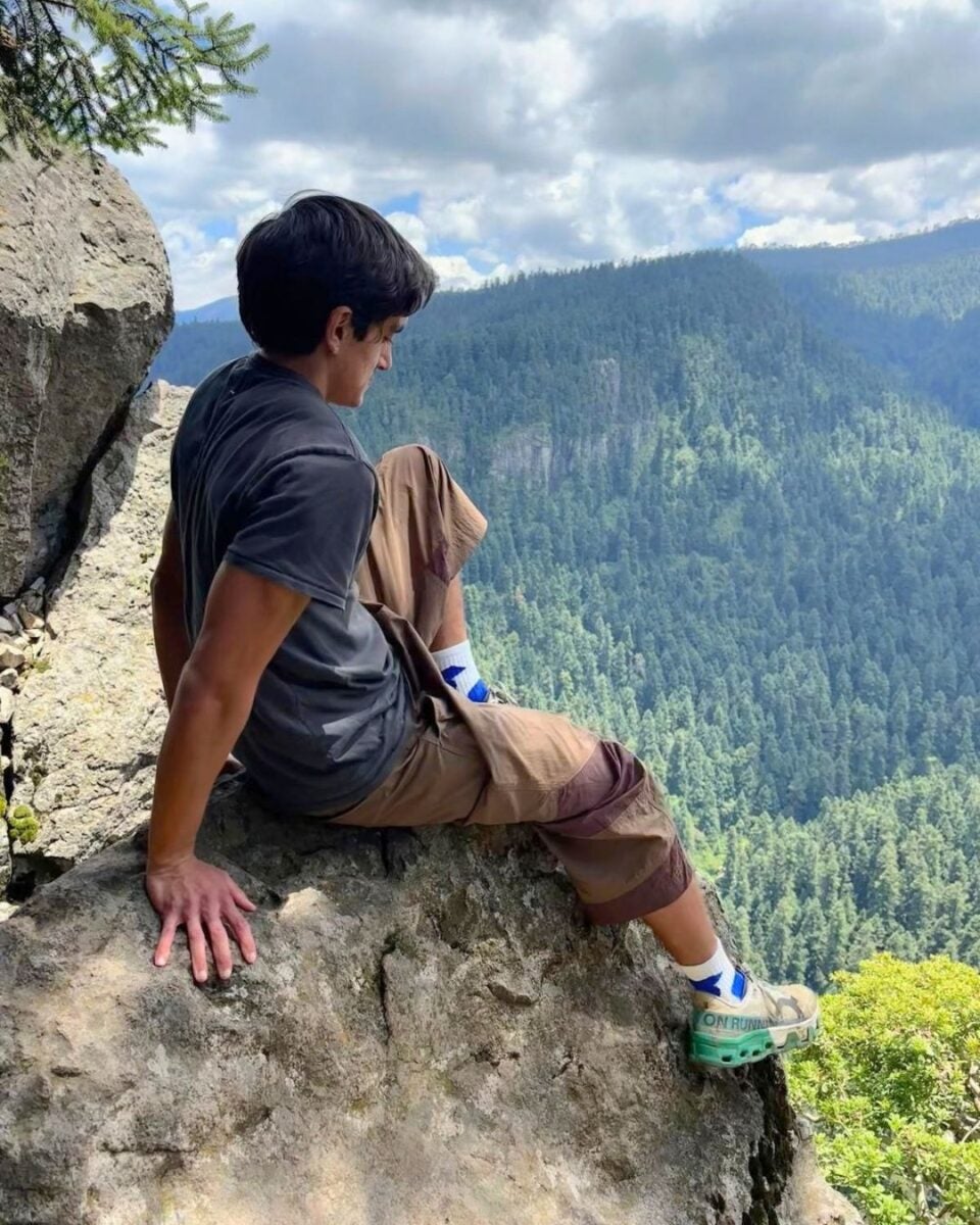 Young man sitting on a rocky cliff edge overlooking a vast forested mountain landscape under a cloudy sky.