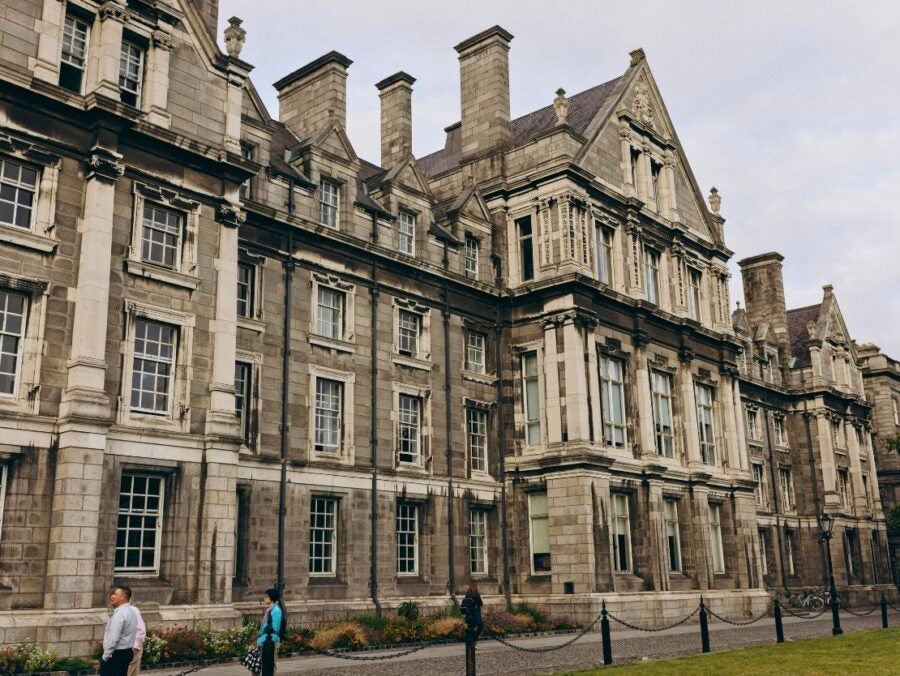 Historic stone building in Dublin, Ireland with tall chimneys and detailed windows under a cloudy sky, with people walking nearby.