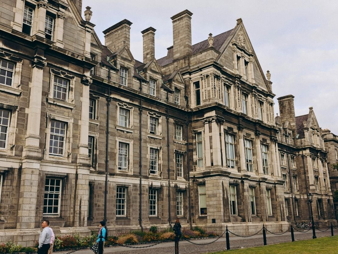 Historic stone building in Dublin, Ireland with tall chimneys and detailed windows under a cloudy sky, with people walking nearby.
