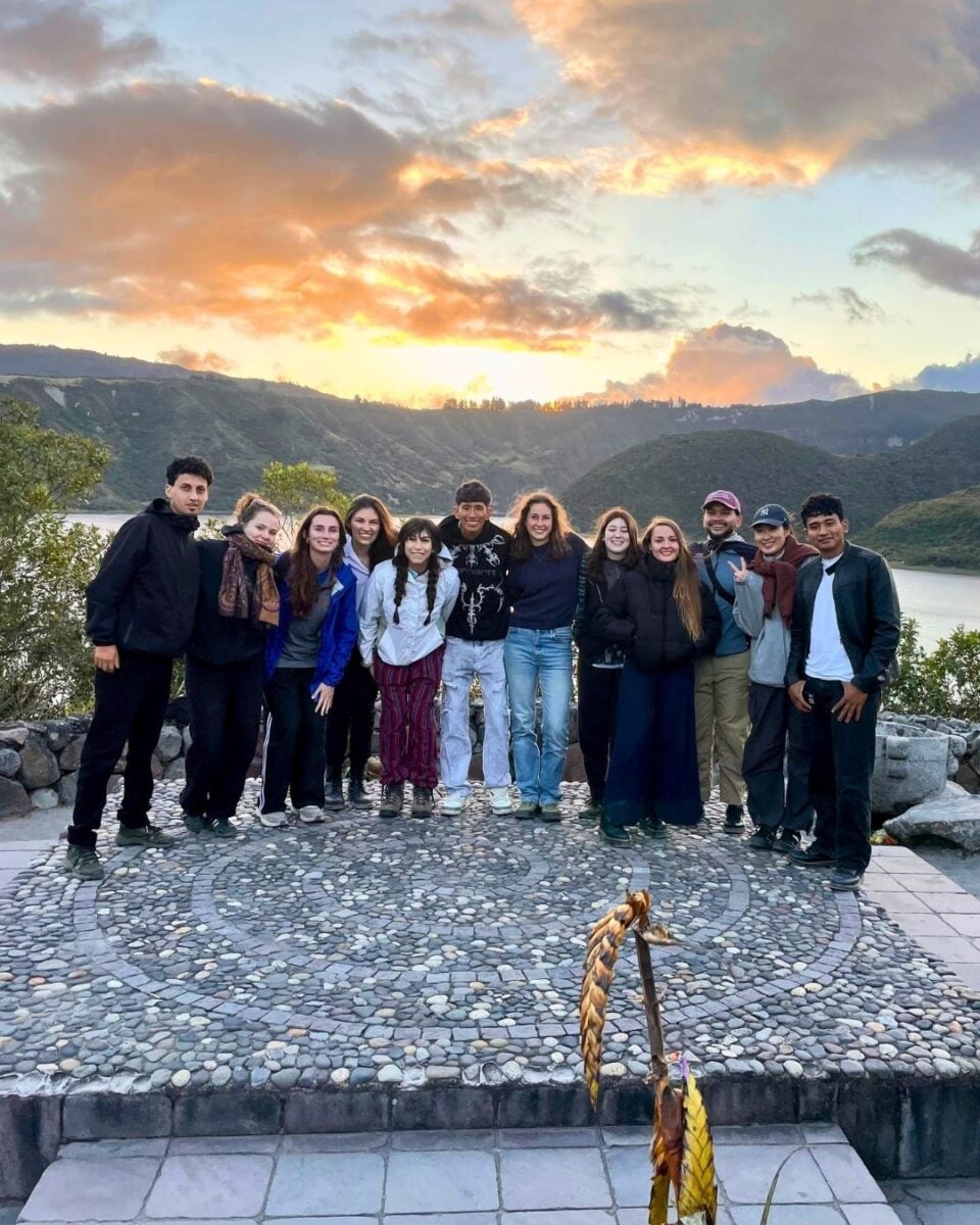 Group of twelve friends standing close together on a stone mosaic platform with a scenic lake and hills at sunset behind them.