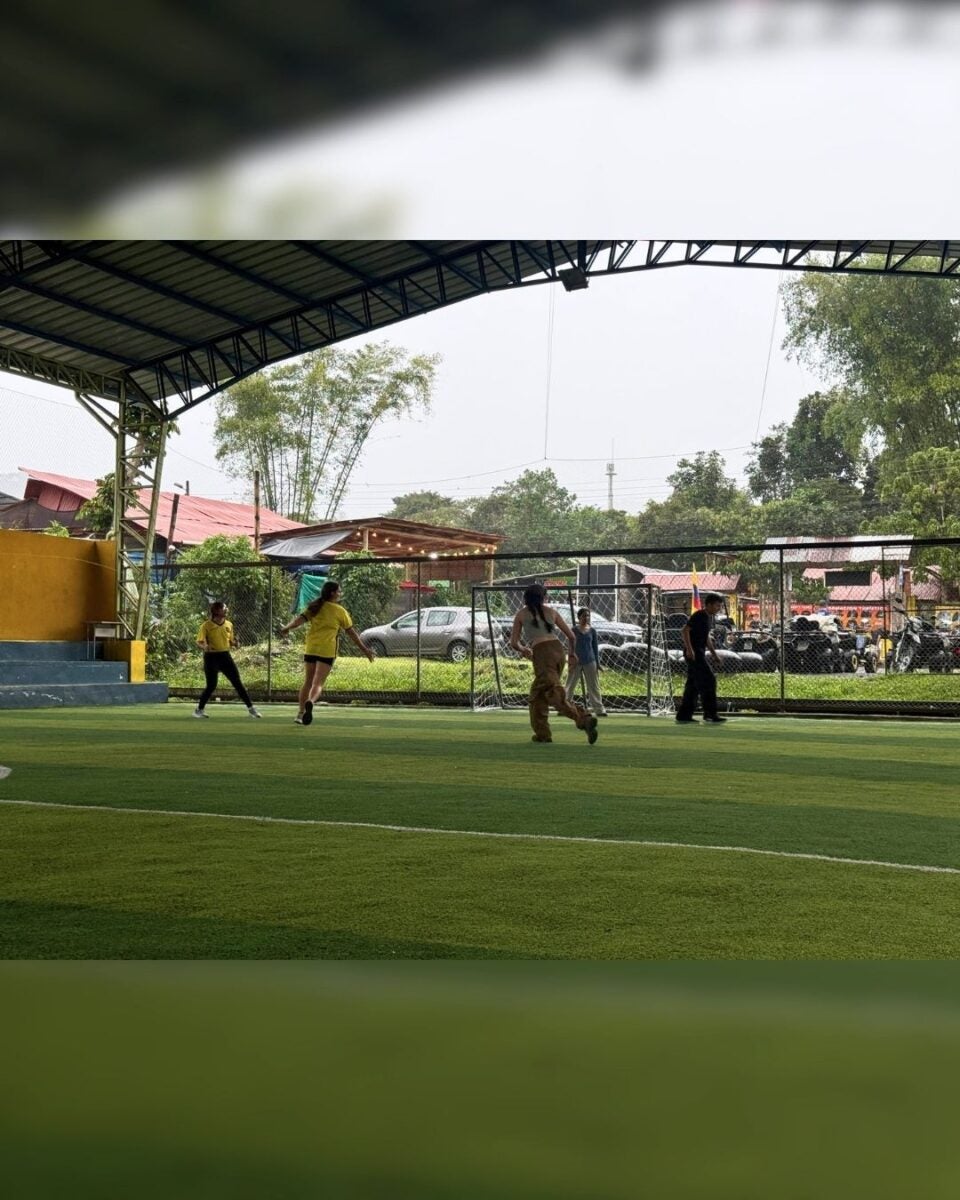 Four people playing soccer on a covered outdoor field with green artificial turf and a metal roof overhead.