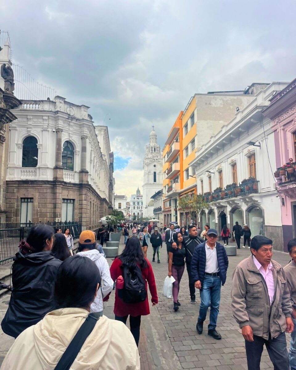 Crowded street scene in a historic city center with colonial-style buildings and a prominent white tower in the background under a cloudy sky.