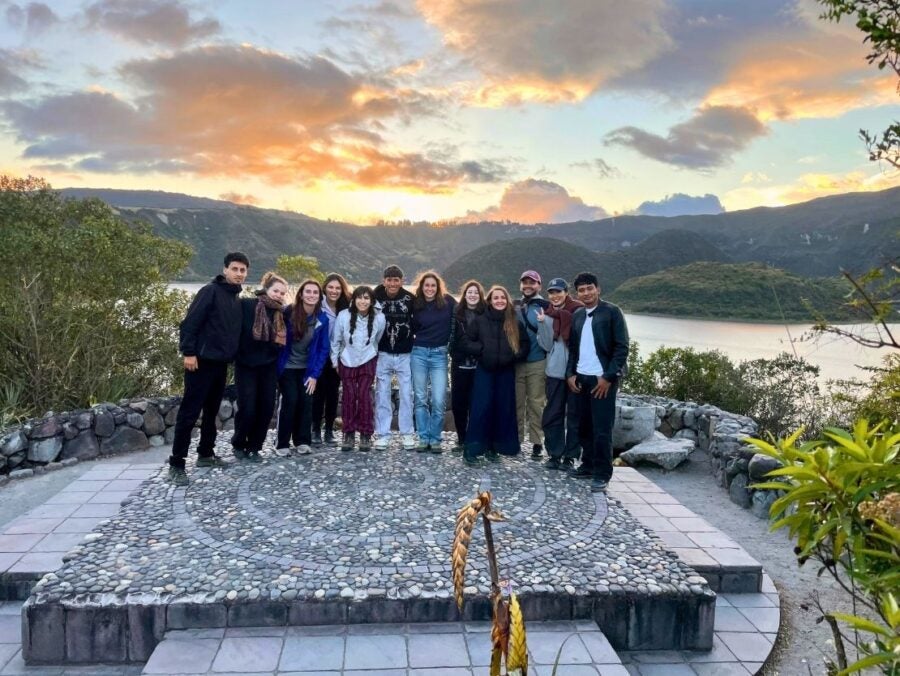 Group of twelve friends standing close together on a stone mosaic platform with a scenic lake and hills at sunset behind them.