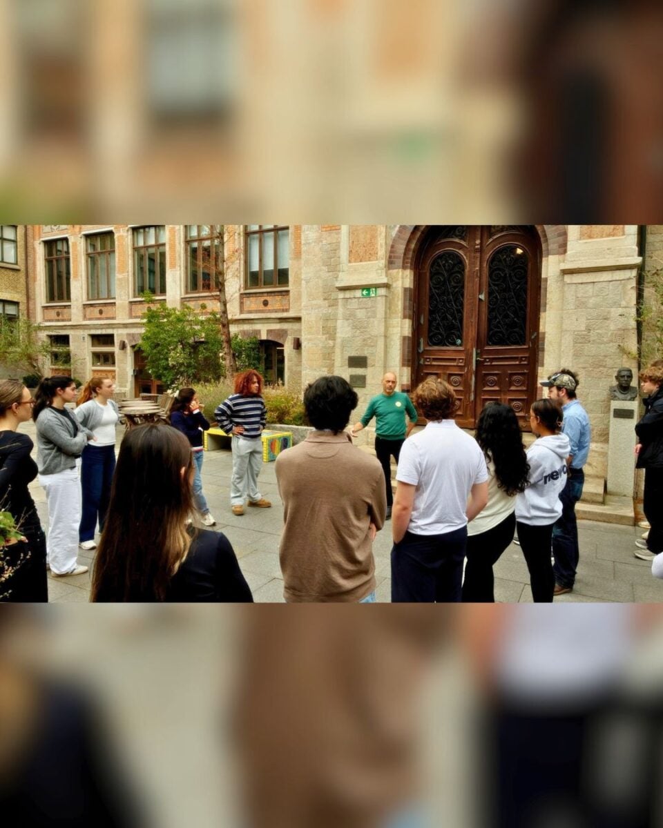 Group of people standing in a semicircle listening to a man speaking outside a historic building with large wooden doors.