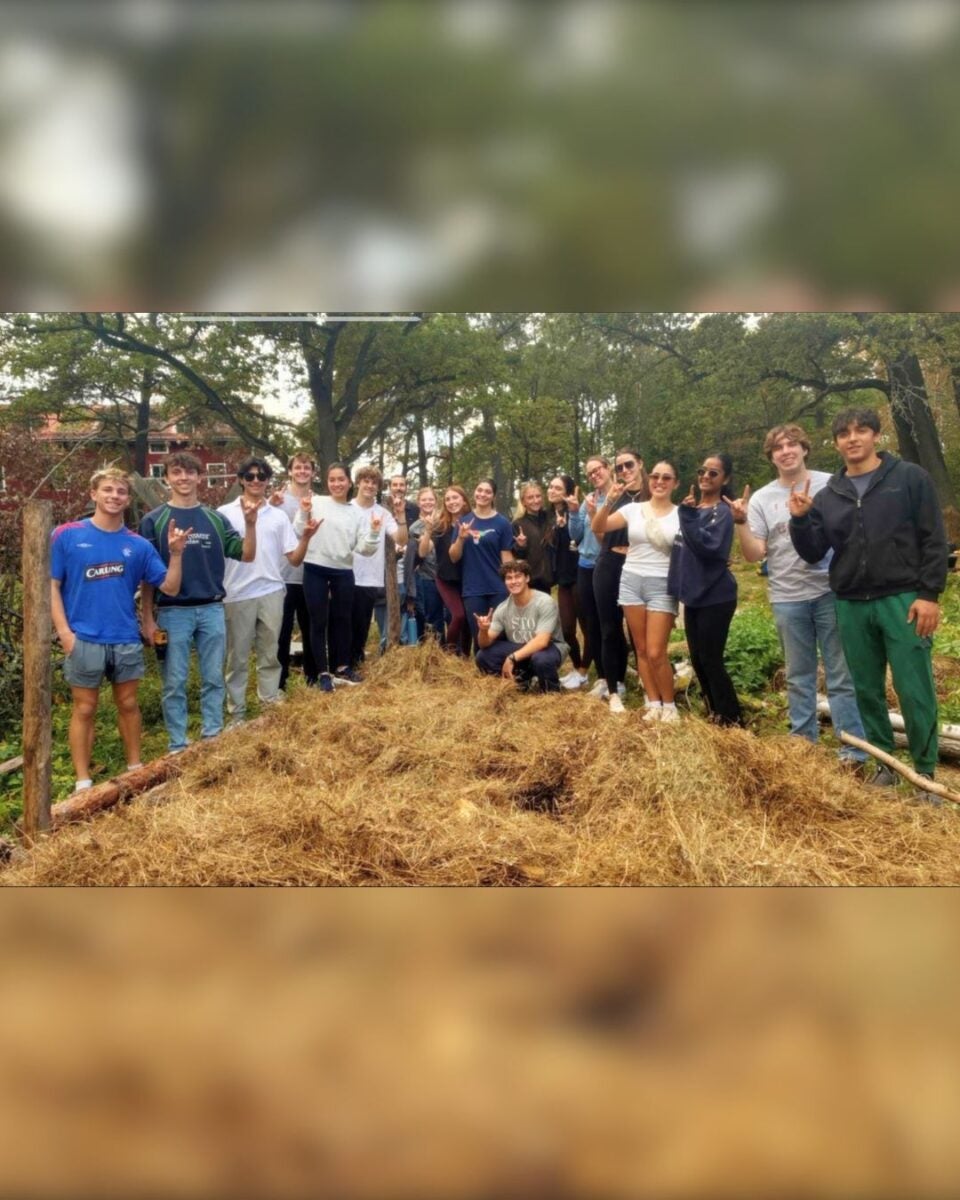 Group of young people standing outdoors around a large pile of straw or mulch in a garden setting, smiling at the camera.