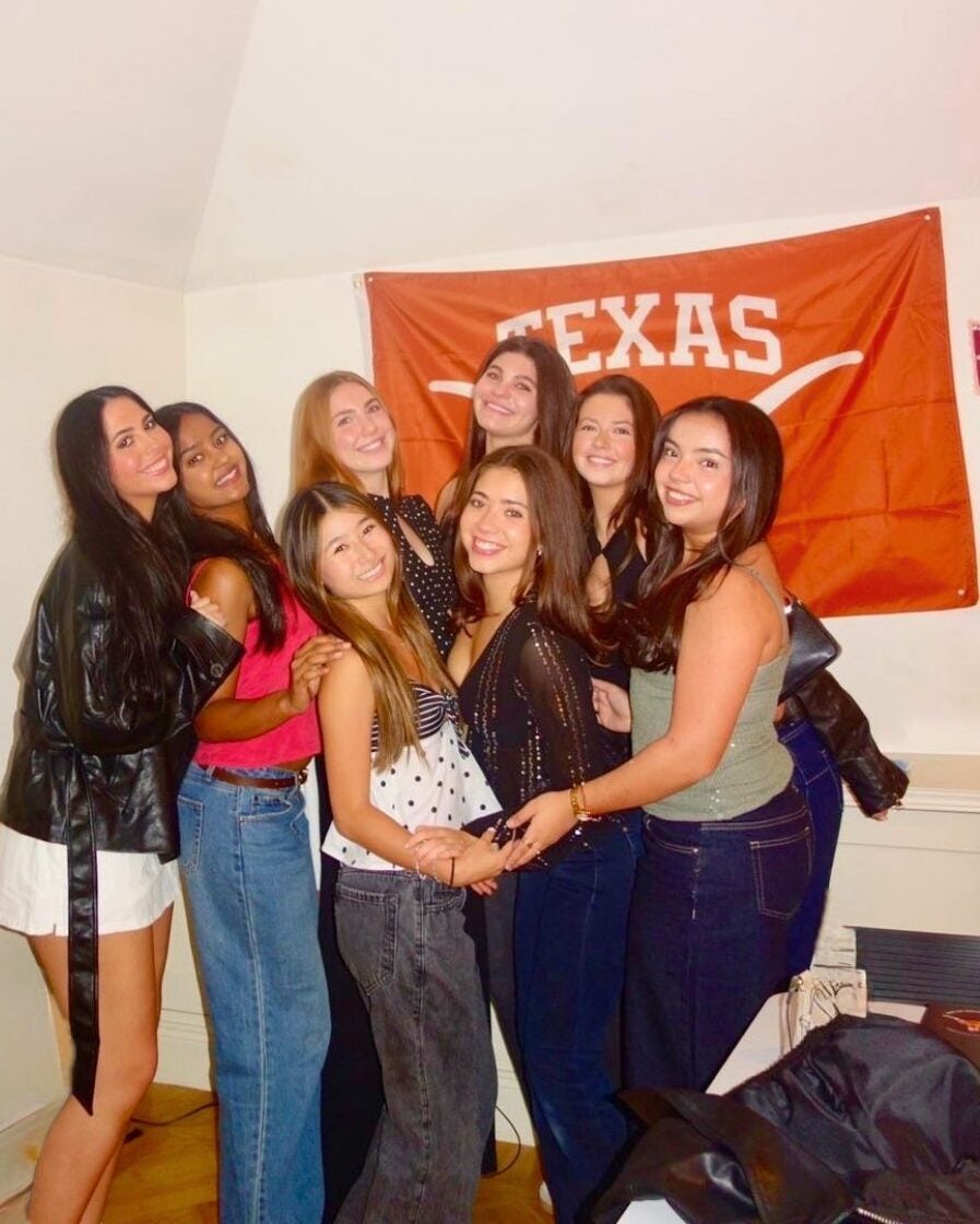 Group of seven young women smiling and posing closely in a room with a Texas flag on the wall behind them.