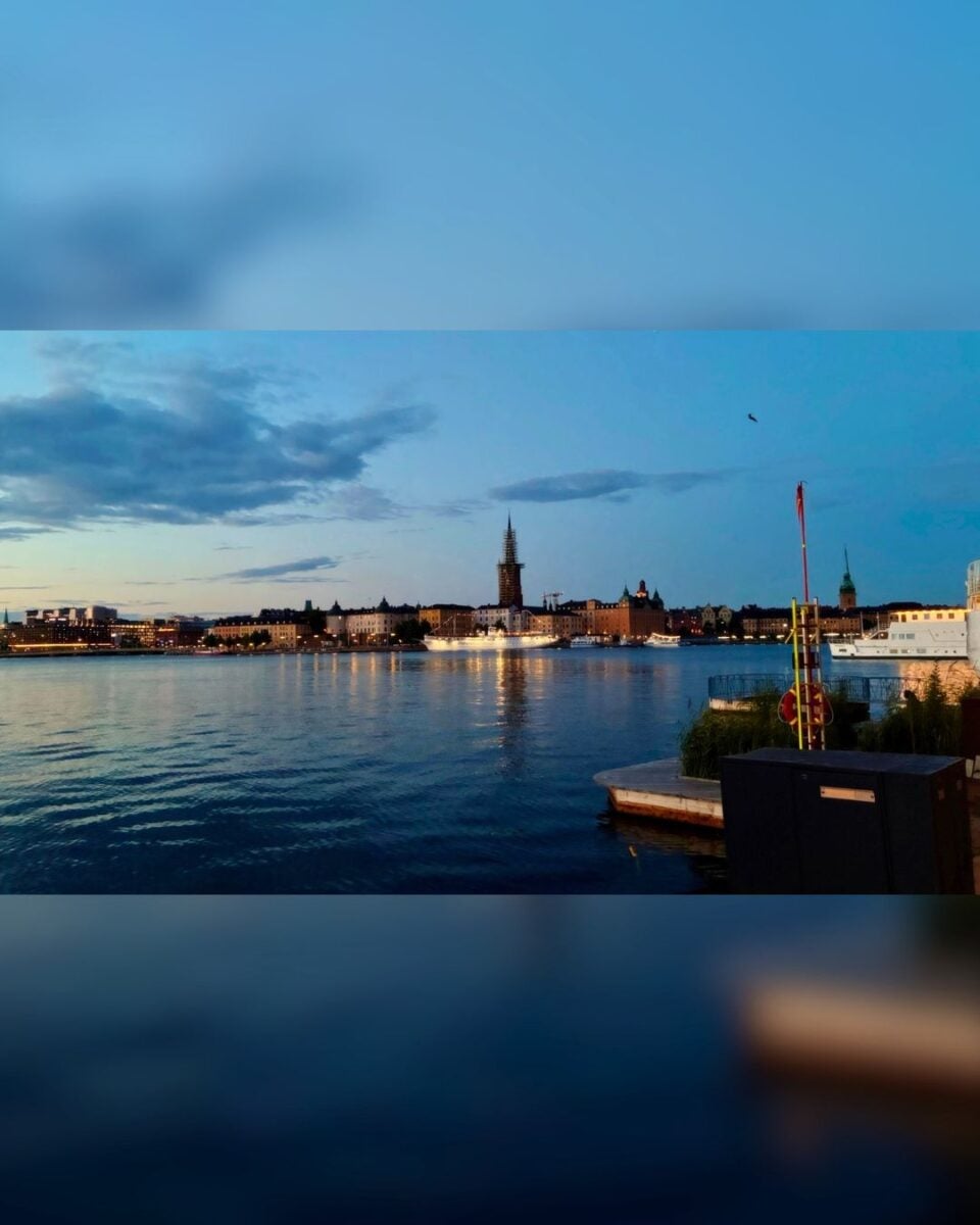 Evening view of a waterfront cityscape with illuminated buildings and a tall spire reflecting on calm water.