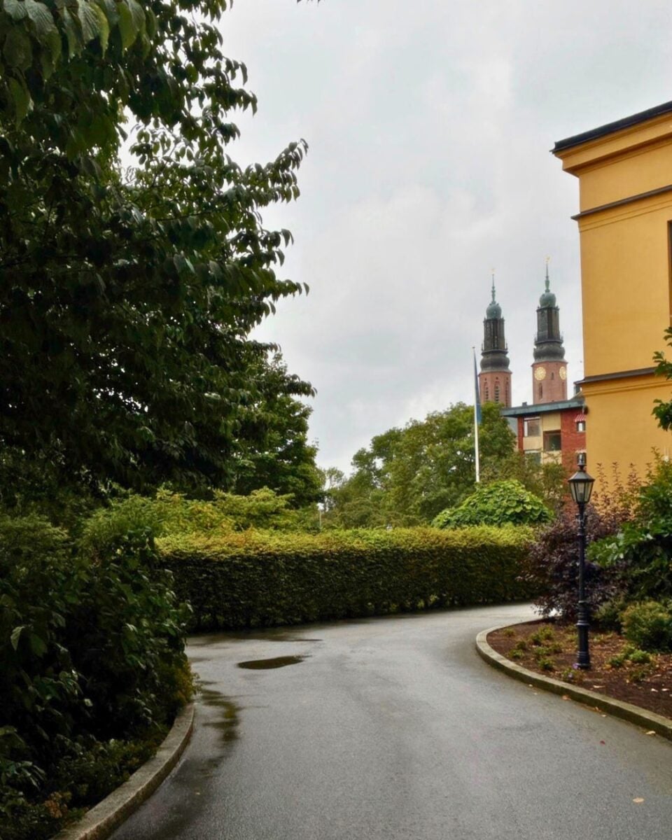 Curved wet pathway bordered by lush green bushes and trees under a cloudy sky with twin church towers in the background.