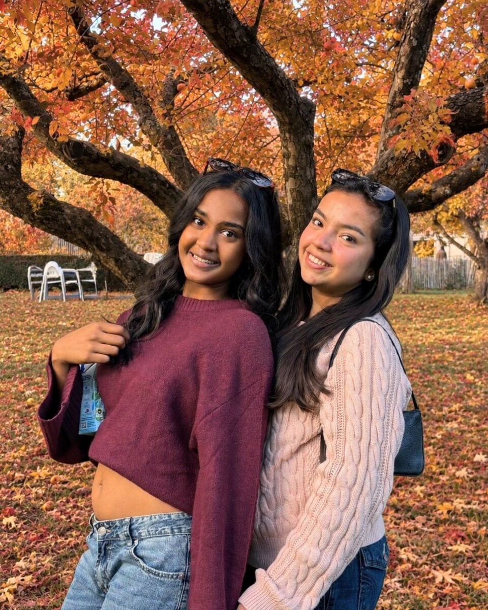 Two young women smiling and posing back-to-back under a large tree with vibrant autumn leaves.