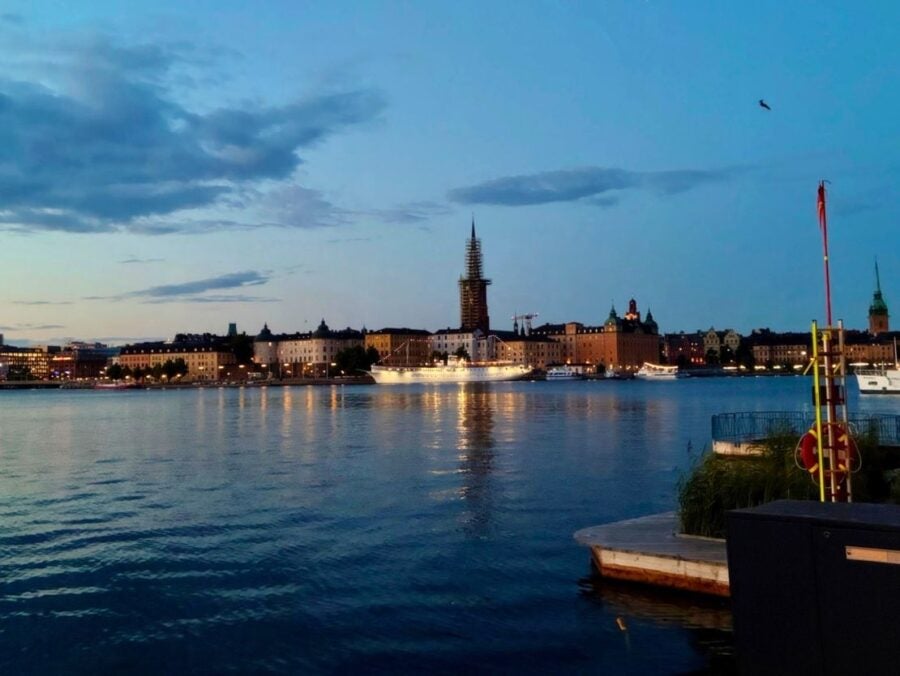Evening view of a waterfront cityscape with illuminated buildings and a tall spire reflecting on calm water.