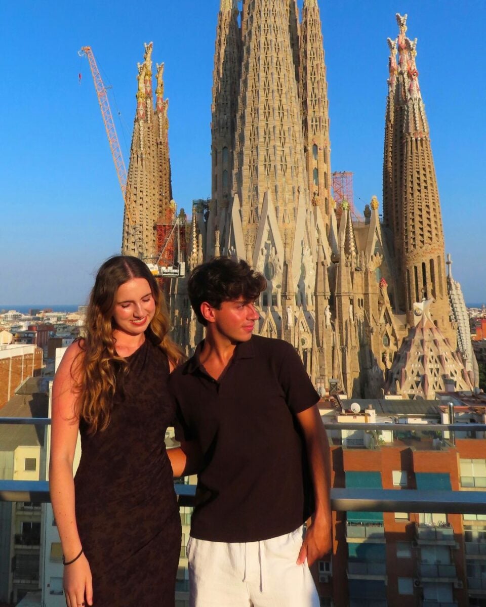 A young woman and a young man posing on a rooftop with the Sagrada Familia basilica towering behind them in bright daylight.