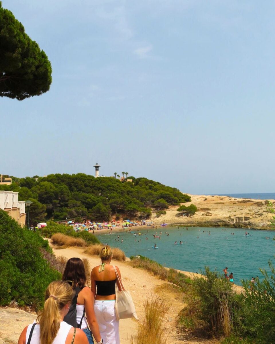 Three women walk along a sandy path toward a crowded beach with turquoise water and a lighthouse on a green hill in the distance.