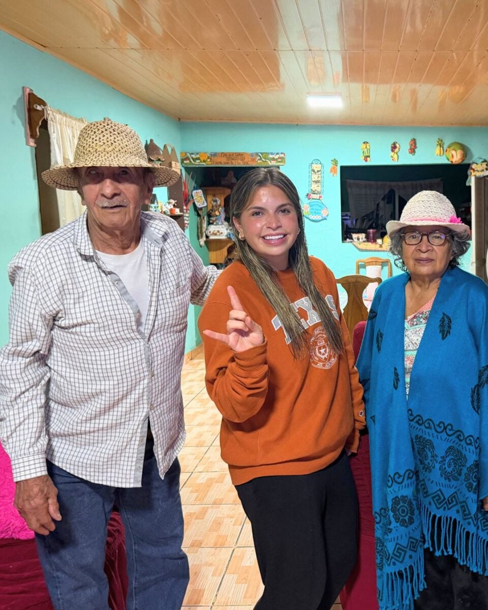 Elderly man and woman wearing hats stand beside a smiling young woman in an orange sweatshirt inside a cozy, decorated room.
