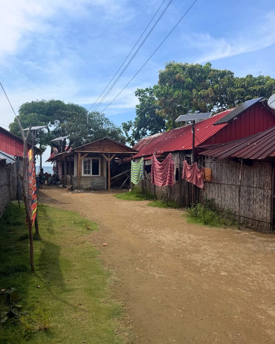 Dirt road lined with houses featuring red metal roofs and clothes hanging on a fence under a clear blue sky.
